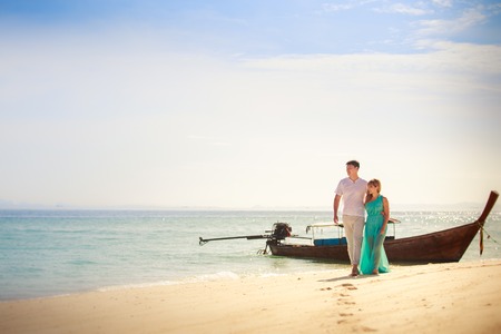 beautiful blonde woman in elegant dress walk with her handsome husband on white sand beach near blue sea with long tail boat on backgroundの写真素材