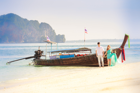 young handsome man and his beautiful blonde girlfriend in blue dress stand near longtail boatの写真素材