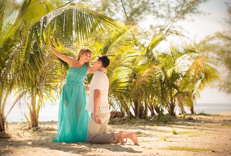 beautiful blonde woman in elegant dress kiss her handsome husband stand in green palm leafsの写真素材