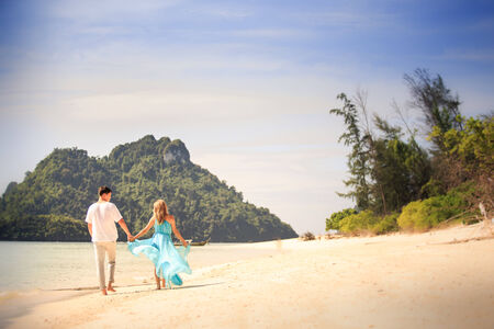 beautiful blonde woman in elegant dress walk with her handsome husband on island beach with green mountain on backgroundの写真素材