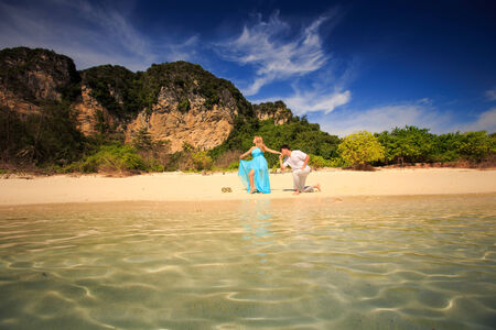 handsome man kiss his beautiful blonde wife in elegant dress on island beach with green mountain on backgroundの写真素材