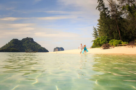 handsome man run with his beautiful blonde wife in elegant dress on island beach with green mountain on backgroundの写真素材