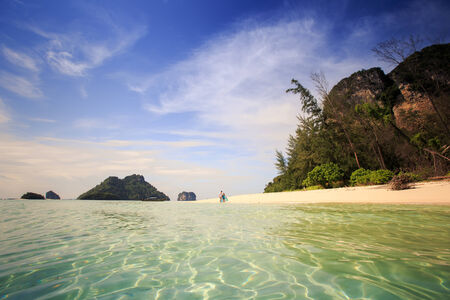 handsome man run with his beautiful blonde wife in elegant dress on island beach with green mountain on backgroundの写真素材