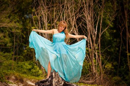 beautiful blonde woman play with her blue dress among green treesの写真素材