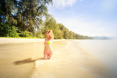 blonde slim girl in swimsuit stands in shallow sea against sun raysの写真素材