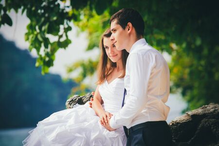 young brunette bride in white wedding dress and handsome groom sit on rock at sand beach against green treesの写真素材