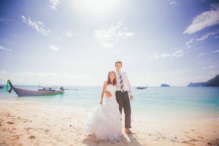 young brunette bride and groom embrace standing on sand beach against azure sea and longtail boatの写真素材