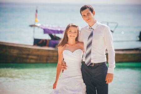 young brunette bride and groom embrace standing on sand beach against azure sea and longtail boatの写真素材