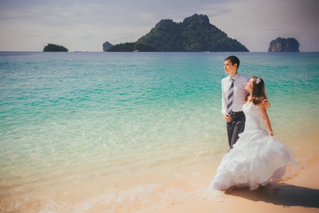 young brunette bride embrace groom walking along sandy beach against azure sea and rocky islandの写真素材