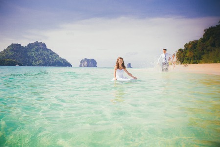 young brunette bride in wedding dress smile playing in shallow azure sea water against rocky islandsの写真素材