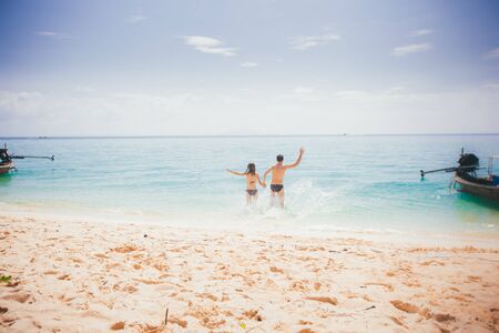 girl and guy run into azure sea waving hands at sand beachの写真素材