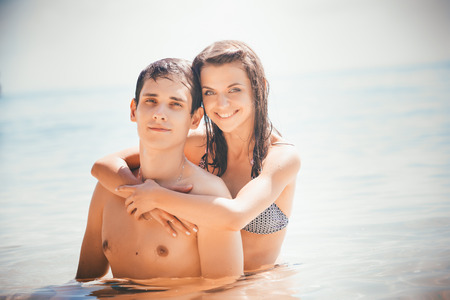 young beautiful girl embrace his boyfriend smiling in shallow water standing against azure sea and boatの写真素材
