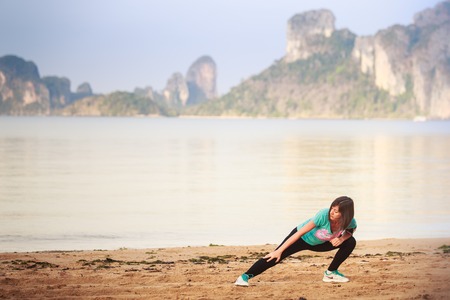 brunette girl does morning exercises on sand beach against cliffy island at dawnの写真素材