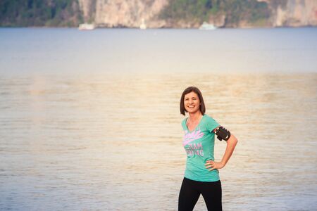 brunette girl in sports clothes walks on sand beach against green island at dawnの写真素材
