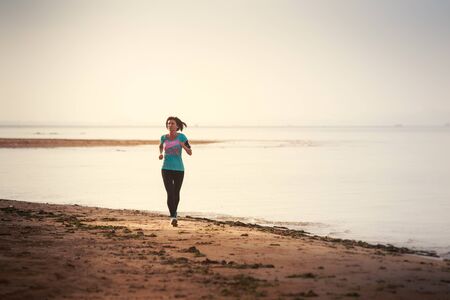 brunette girl in sports clothes runs on sand beach at low tide against sea at dawnの写真素材