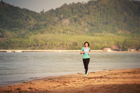 brunette girl in sports clothes runs from distance on sand beach at low tide against sea at dawnの写真素材