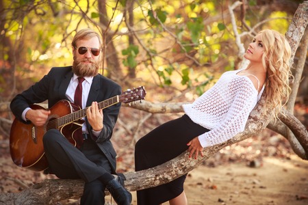 bearded guitarist in black suit and blonde girl  in purple dress sit on branch of tropical treeの写真素材