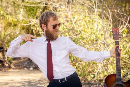bearded guy in white shirt pose with guitar against treeの写真素材