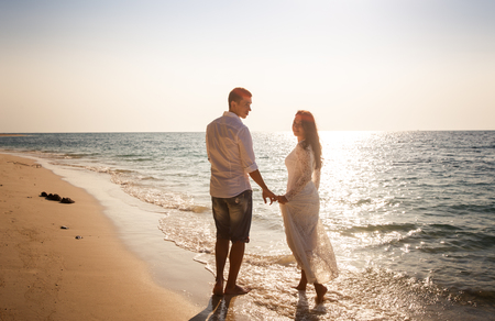 brunette bride and groom barefoot at edge of transparent water against backlight of sun raysの写真素材