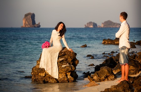 brunette bride sits on rock in sea and handsome groom stands on beach at sunriseの写真素材