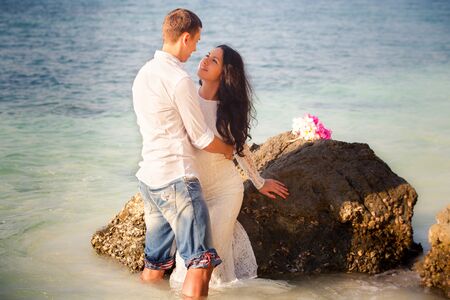 brunette bride and groom hug at rock at beach at sunriseの写真素材