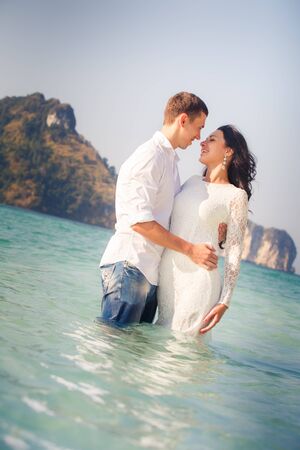 brunette bride and groom hug half in sea water at beach at sunriseの写真素材
