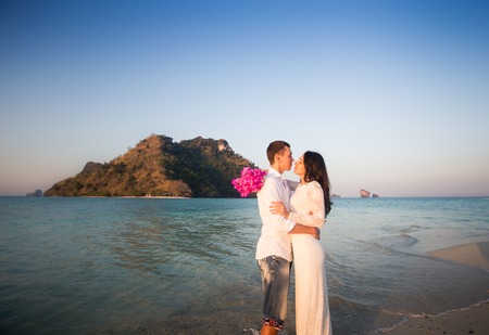 brunette bride with pink bouquet and handsome groom hug and kiss at sand beach against cliffの写真素材