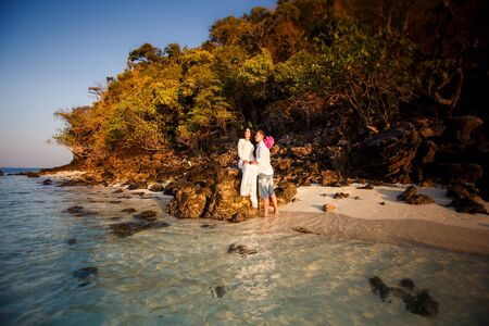 brunette bride and handsome groom at beach against rocks at sunriseの写真素材