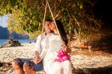 brunette bride with pink bouquet and handsome groom are on rope swing on beach of tropical islandの写真素材