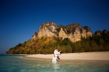 groom holds bride in arms barefoot at edge of transparent water against cliffsの写真素材