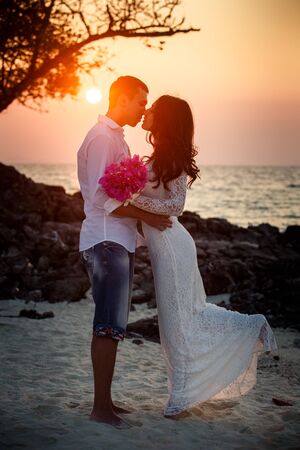 brunette bride and handsome groom barefoot hug at sand beach at sunriseの写真素材
