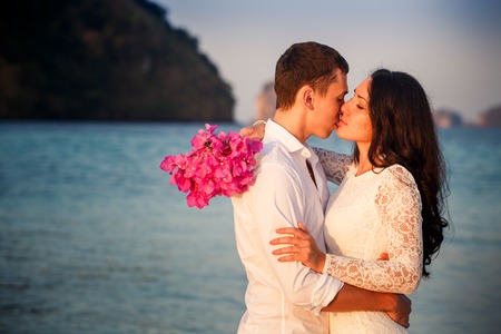brunette bride and handsome groom barefoot kiss at sand beach at dawnの写真素材