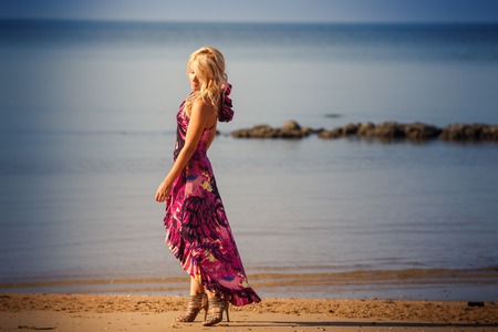 blonde girl in purple dress walks on sandy beach against azure sea at sunriseの写真素材