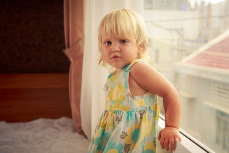 little blonde girl in colourful dress stands by window on sofa side-viewの写真素材