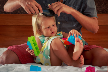 father brushes hair of little blonde daughter sitting near on sofa with toy constructorの写真素材