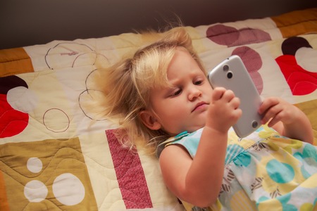 pretty blonde two years old girl plays with smartphone lay on colored couchの写真素材