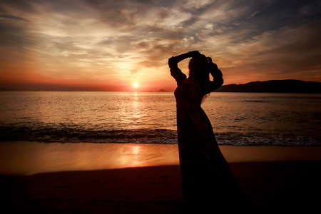 silhouette of slim longhaired girl in long dress with hands up on beach at background of rising over sea sun and cloudsの写真素材
