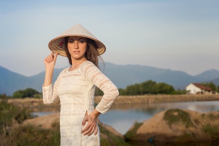 slim blonde girl in Vietnamese national white long dress and hat smiles against lakes country house and mountainsの写真素材