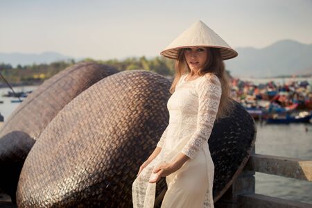slim blonde girl in Vietnamese national white long dress touches hat by barrier of embankment against boats mountainsの写真素材
