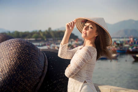slim blonde girl in Vietnamese national white long dress touches hat by barrier of embankment against boats mountainsの写真素材
