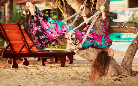 slim brunette longhaired girl in long and big red hat swings on rope swing by tropical tree at defocused backgroundの写真素材
