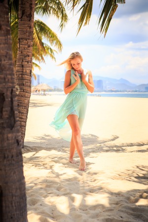 European slim blonde girl barefoot in light azure transparent frock stands near palms with hands on breast on beachの写真素材