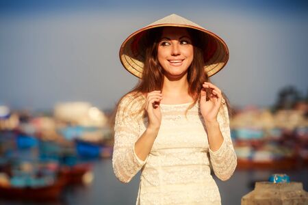 slim blonde girl closeup in Vietnamese national white dress and hat leans on barrier against defocused boatsの写真素材