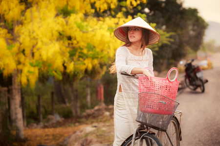 slim blonde girl in Vietnamese long dress and hat closeup looks sideward by bike basket against defocused yellow plantsの写真素材