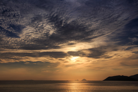 panorama of sunrise from behind golden cumulus clouds from beach above sea and island with sunlight reflection pathの写真素材