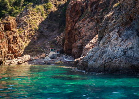 green and brown rocky wall of island above azure sea with two small buildings in gorge of bayの写真素材