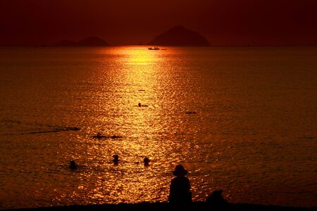 people swim in sea among sun reflection against red sky and islands at sunriseの写真素材
