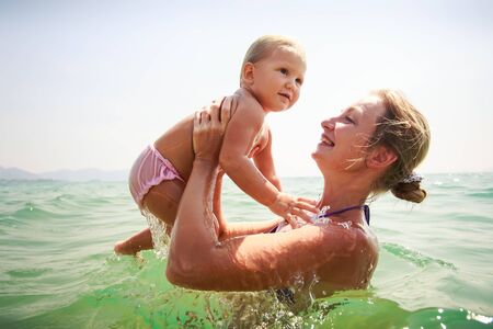 closeup mother and small blonde daughter bathe in transparent azure sea smile against skyの写真素材