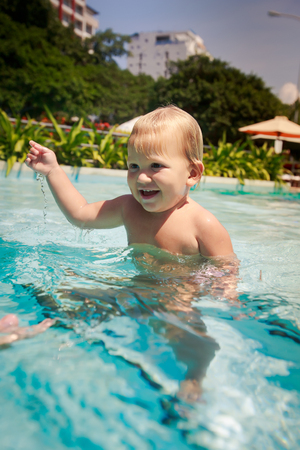 closeup portrait small blonde girl stands smiles and looks into water in hotel swimming pool against tropical plantsの写真素材