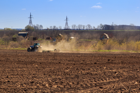panorama of tractor operating in field at background of aerial high-voltage line and small houses in early springの写真素材